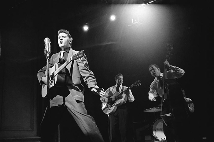 A picture of Elvis Presley on stage singing while holding his acoustic guitar in one hand and two band members visible in the background playing their instruments.